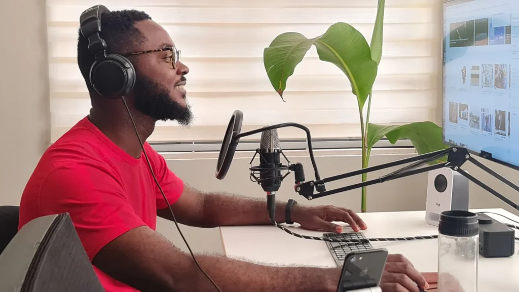 A man in a red shirt wears headphones and speaks into a microphone at a bright home-office desk.