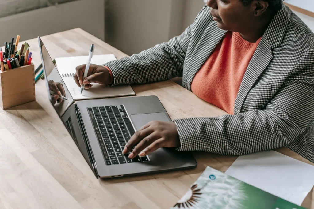 Woman in a checked blazer writing in a notebook beside an open laptop at a wooden desk.