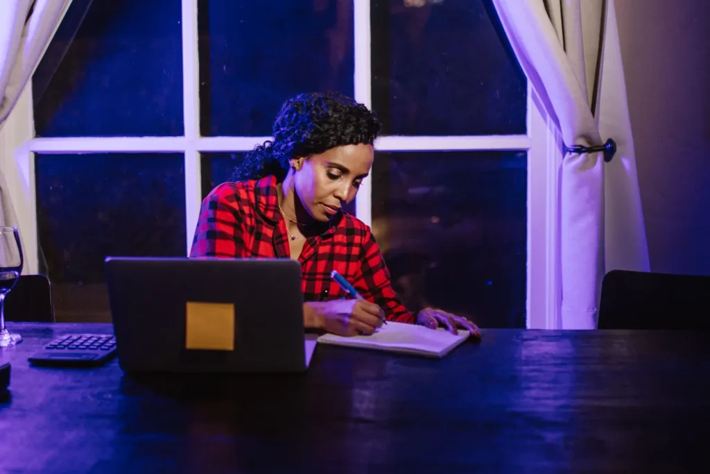 A woman writes in a notebook beside an open laptop at a table illuminated by blue and violet light.