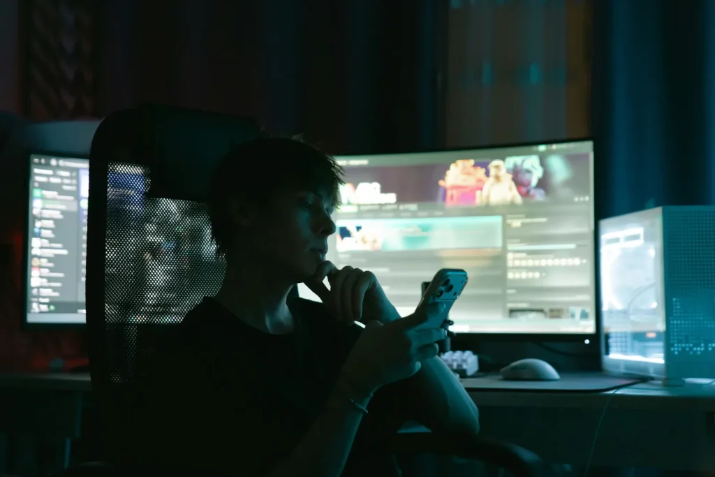 A person looks at a smartphone while sitting in front of glowing desktop monitors in a dark room.