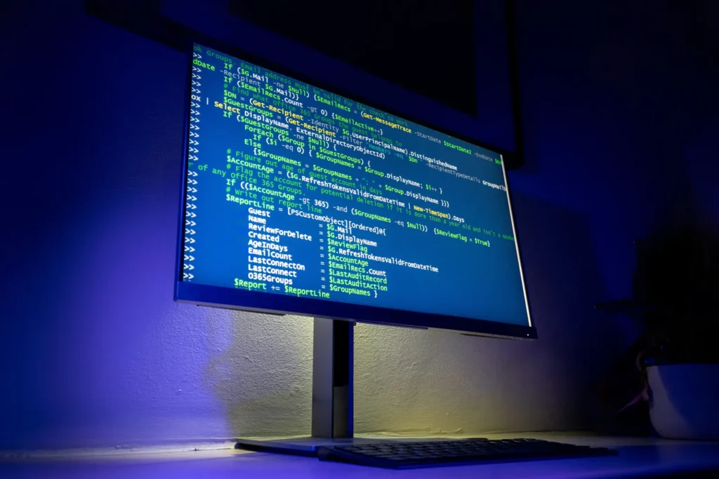 A desktop monitor displays lines of code above a keyboard in a dark room with blue lighting.