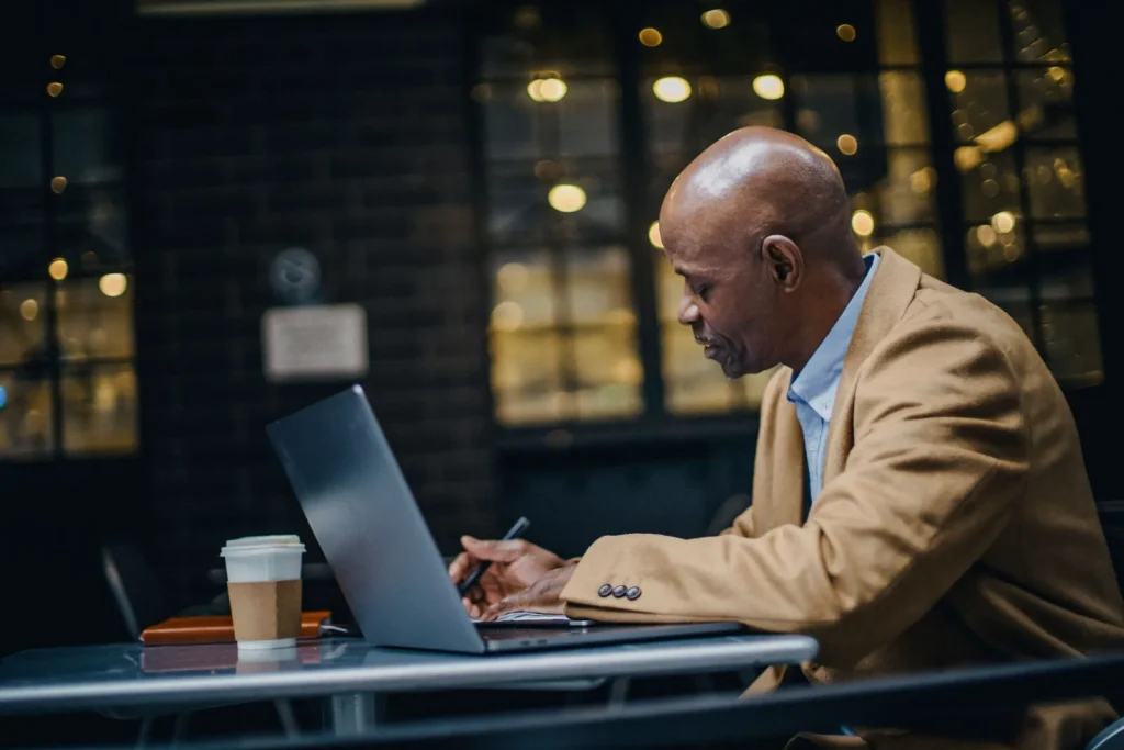 A man in a tan jacket writes notes beside an open laptop and a takeaway coffee at a cafe table.