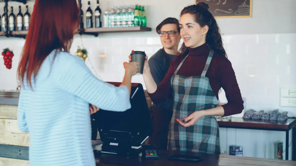 A smiling barista hands a takeaway coffee to a customer across a cafe counter.