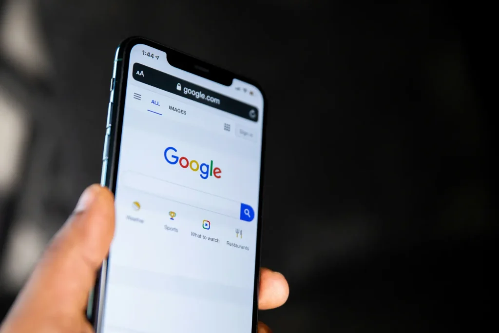 A hand holds a smartphone showing the Google search homepage against a dark background.
