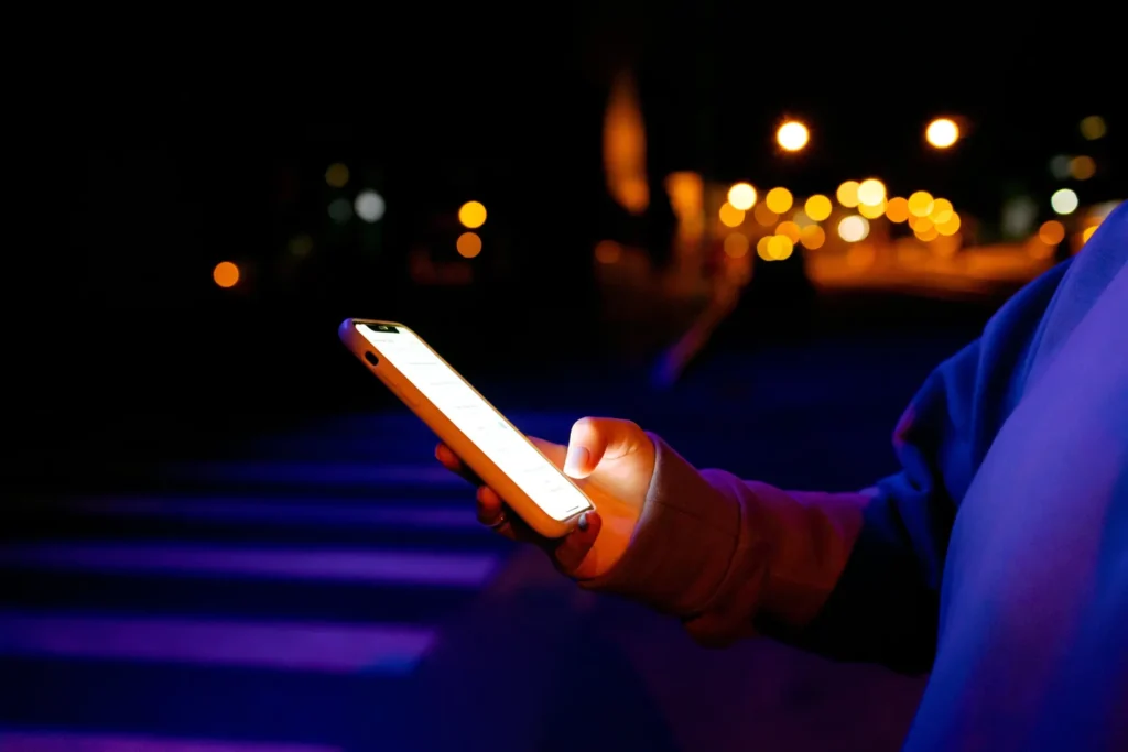 A person holds an illuminated smartphone outdoors at night, with blue light on their sleeve and amber city lights behind them.