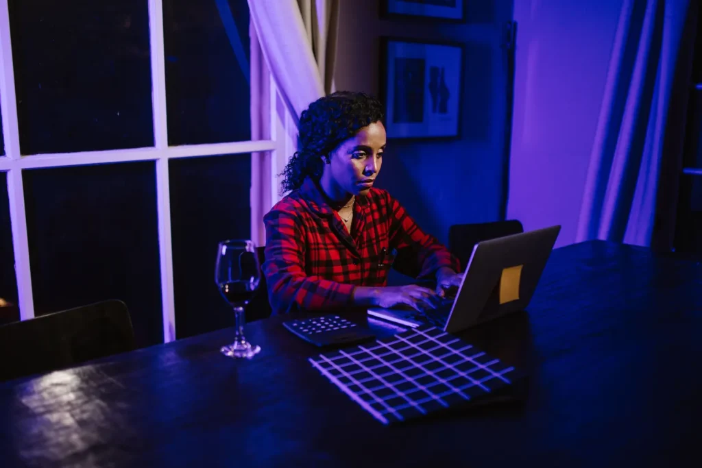 A woman in a red checked shirt works at a laptop beside a window in a room lit with blue and violet light.